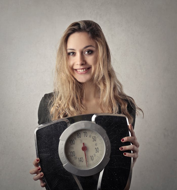 hero-img-01 Young woman with blond hair smiling, holding a vintage bathroom scale indoors.